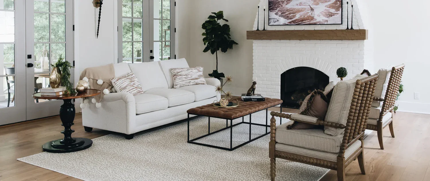 A modern living room with a white sofa, two patterned armchairs, a brown ottoman, and a side table with decorative items. There is a white brick fireplace with a wooden mantel, a large windowed door to the left, and a flat-screen TV mounted above the