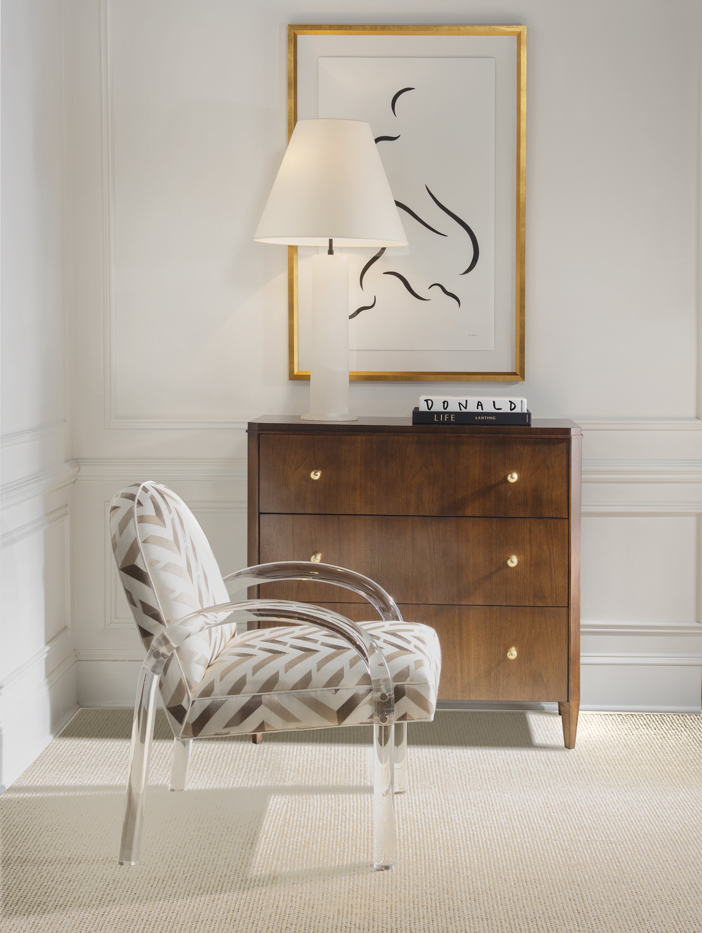 A white and clear acrylic chair with a beige patterned fabric cushion in front of a brown wooden dresser with three drawers, each with a small round golden knob. On top of the dresser is a white lamp with a white shade, and two stacked books labeled "DONALD LIFE" and "LANTING." Behind the lamp is a large framed line art portrait of a woman's face outlined with black strokes, hanging on a white wall with decorative wainscoting.