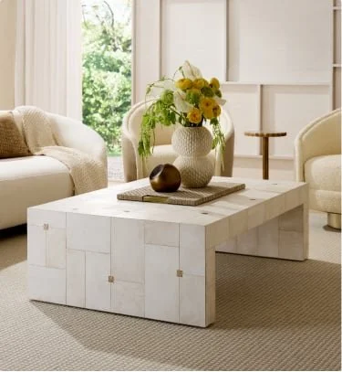 Living room with white wooden coffee table, a vase of yellow and white flowers, and a brown decorative object. Sofa and armchair in background, with large windows letting in natural light.