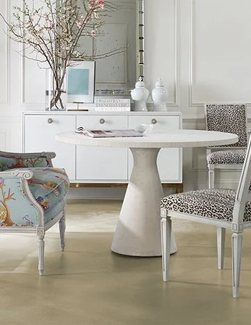 Dining room with a white round table, a patterned chair on one side, a floral armchair on the other, and a white sideboard with decorative objects and artwork in the background.