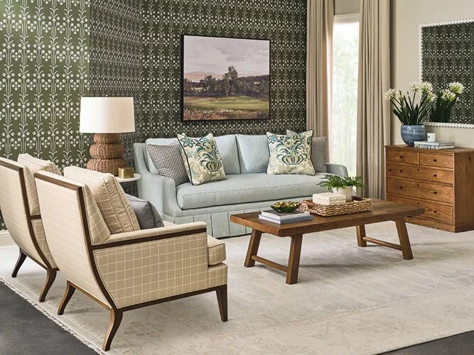 Living room with patterned dark green wallpaper, light gray sofa with decorative pillows, two beige armchairs, wooden coffee table, sideboard, floral arrangement, lamp, and painting on the wall.
