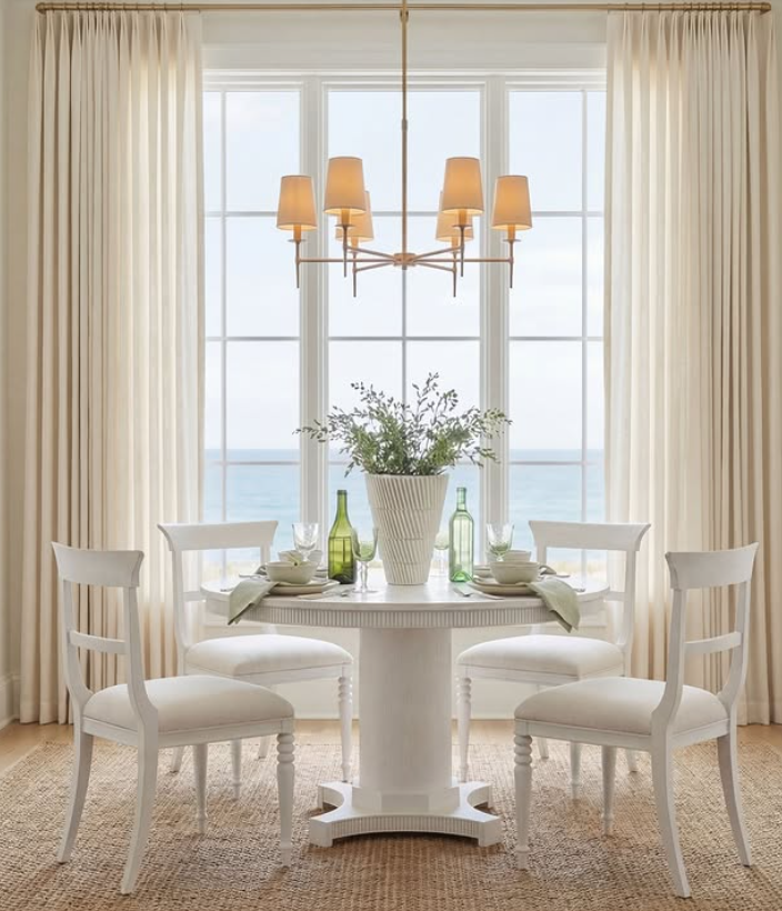 Dining area with a round white table set with cups, plates, and green bottles. Four white chairs surround the table. A large potted plant is in the center. Behind, tall windows with sheer curtains and a view of the sea. A chandelier with multiple bei