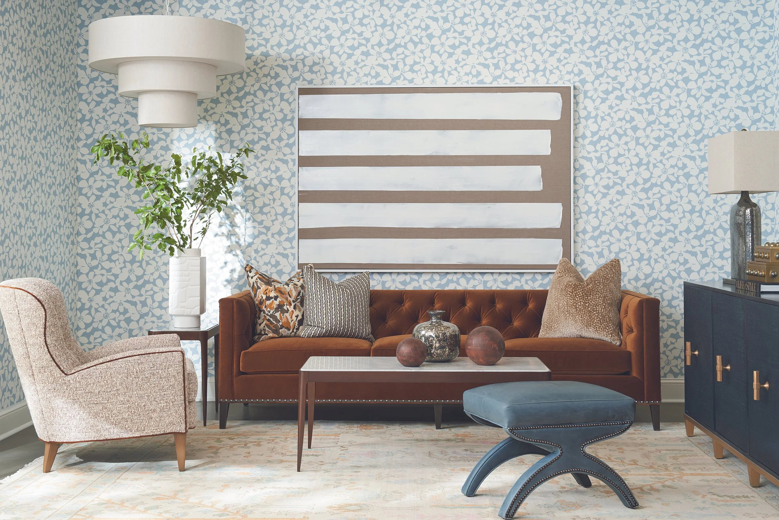 Living room with patterned blue wallpaper, a brown velvet sofa with decorative pillows, a small white coffee table with decorative objects, a beige armchair, a white plant in a vase, a large white and wood wall art piece, and lamps on side tables.