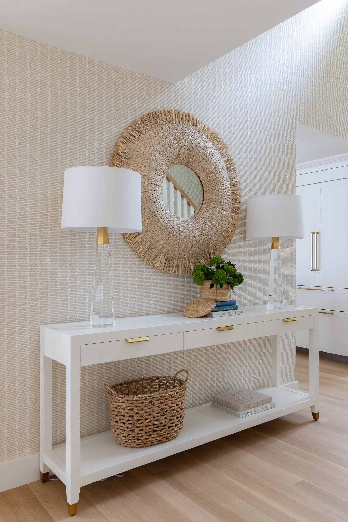 Entryway with a white console table, wicker basket, green plant, two white lamps, round woven mirror, and neutral patterned wallpaper.