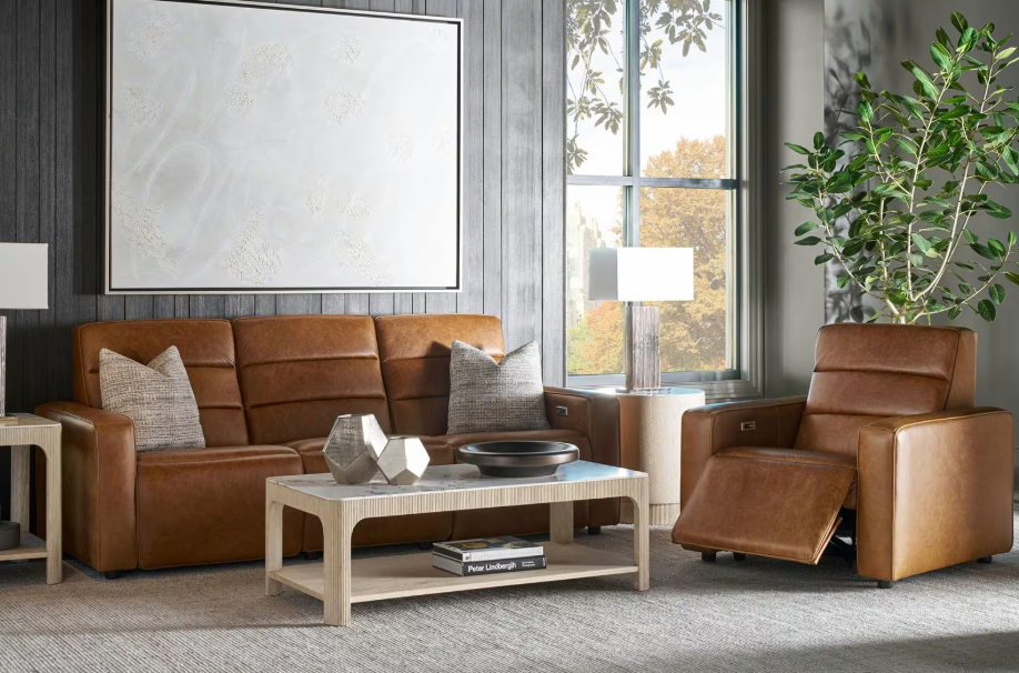 Living room with brown leather sofa and armchair, side table with lamp, modern coffee table with decorative items, large window with view of trees, potted plant, and minimalist wall art.