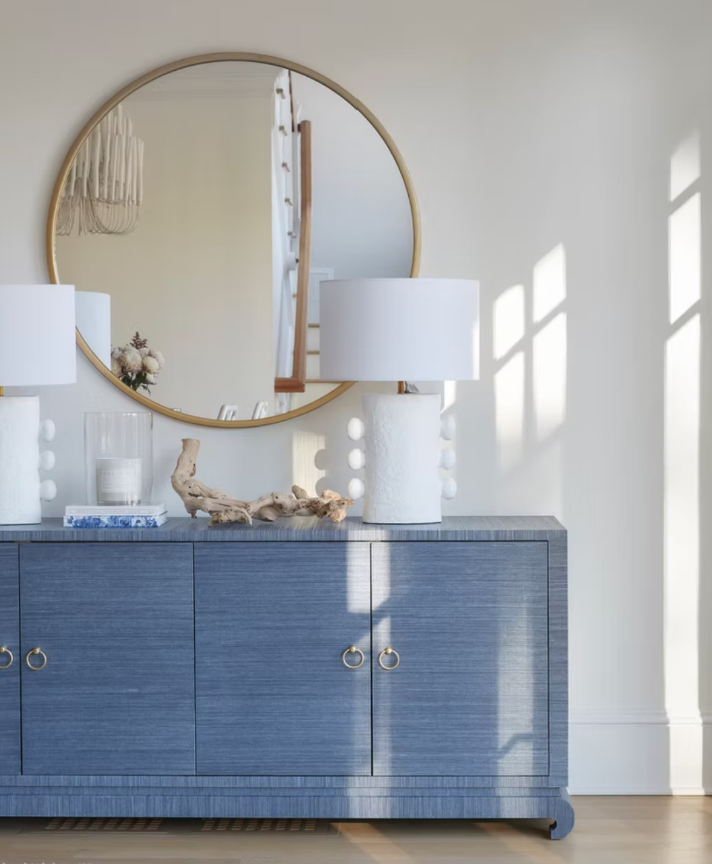 A blue wooden sideboard with two large white table lamps on top, a mirror hanging on the wall, a small floral arrangement, and decorative driftwood, with sunlight casting shadows on the white wall.