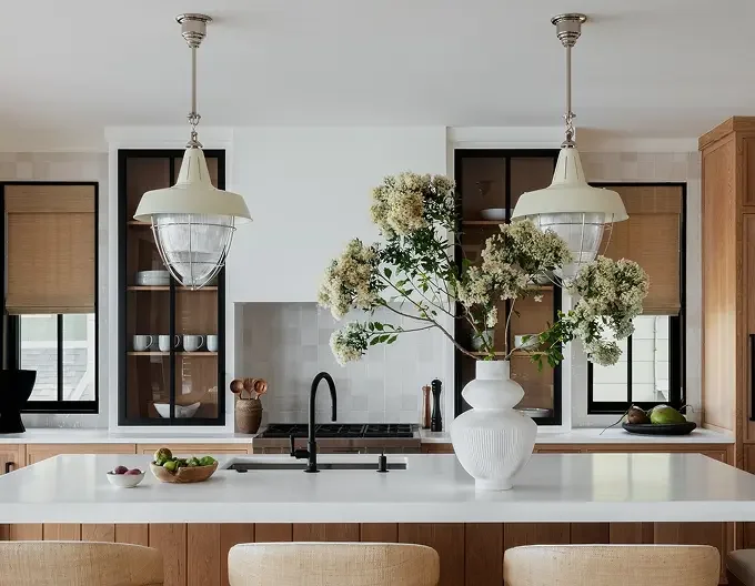 Modern kitchen with white island, black fixtures, and wooden cabinets, featuring a large white vase with flowers, pendant lighting, and windows with bamboo shades.