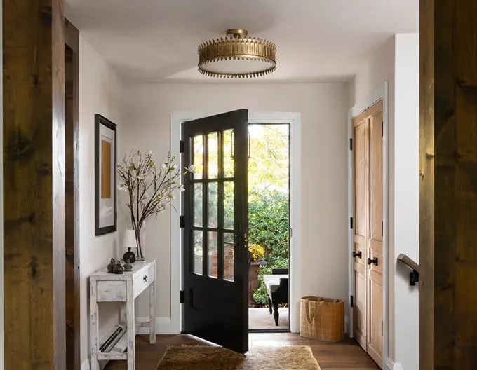 Entrance hallway with open black front door leading to a porch with greenery and a chair, white console table with plants and decorative items, framed artwork on the wall, wooden closet door, and a woven basket, modern light fixture on the ceiling.