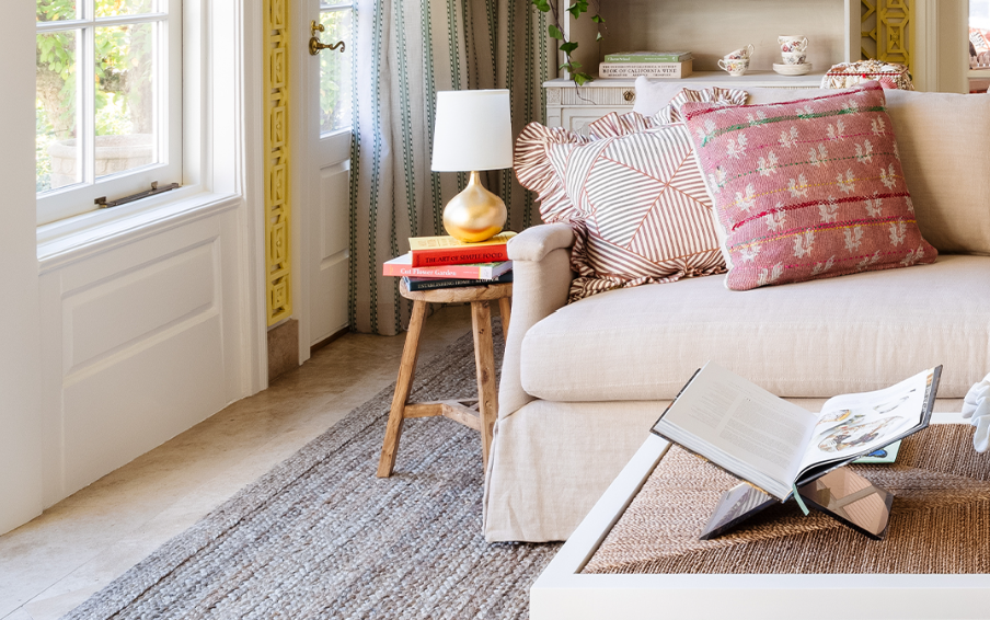 Living room corner with a beige sofa, decorative pillows, a wooden side table with a lamp and books, and a window with patterned curtains.