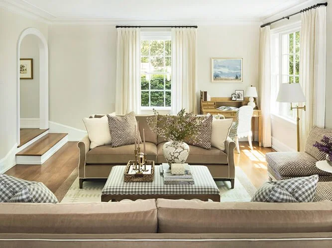 Living room with beige sofas, checkered and plain pillows, a coffee table with a decorative vase and books, a wooden writing desk with a white chair, large windows with cream curtains, hardwood floors, and framed artwork on the wall.