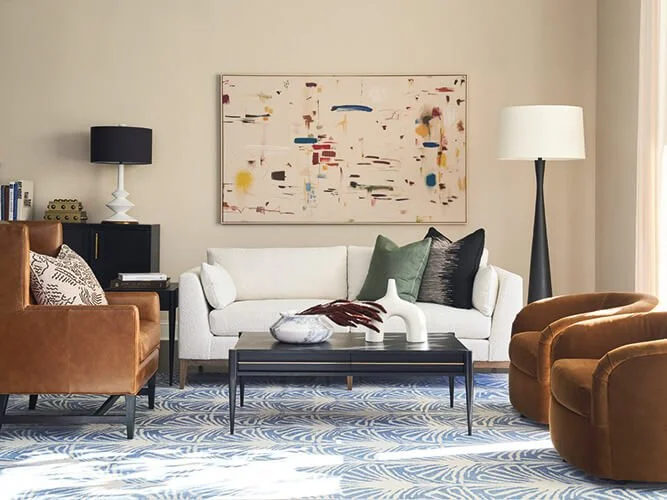 Living room with a white sofa, two green cushions, and a dark cushion, brown armchairs, a black coffee table with a white decorative object and a bowl, a large abstract painting, a black table lamp, and a white floor lamp, with a blue patterned rug.