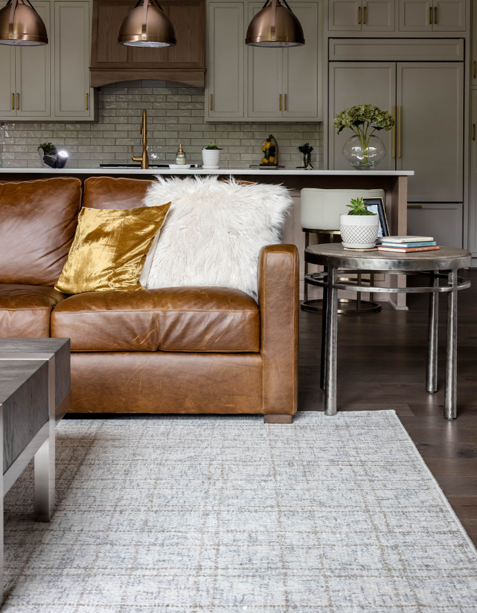 Living room with a brown leather sofa, gold and white pillows, a rustic side table with a potted plant and stacked books, and a kitchen in the background with white cabinets, a white countertop, and decorative items.