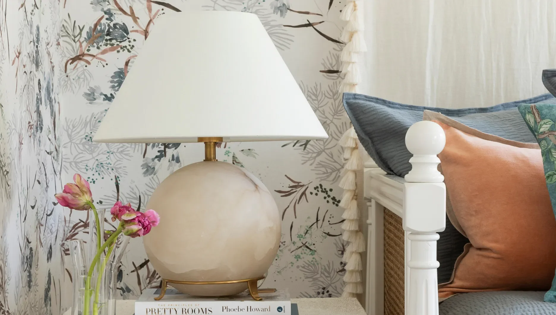 A bedside table holding a book titled 'The Principles of Pretty Rooms' by Phoebe Howard, topped with a textured white ceramic table lamp with a white shade, and a small vase with pink flowers. The background shows a bed with a white wooden post and a
