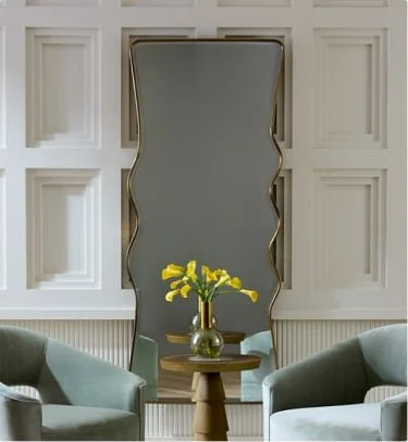 A mirror with a gold frame on a wall behind a small wooden table with a gold vase containing yellow calla lilies, flanked by two light green armchairs.