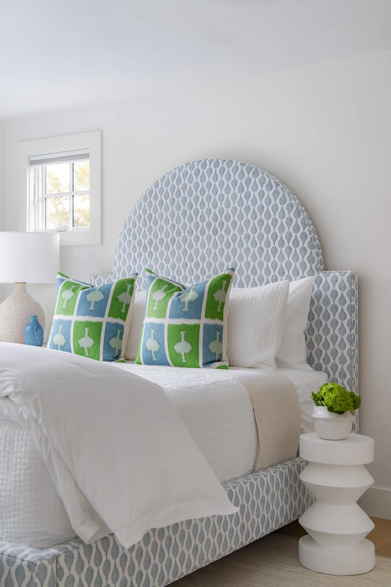 A neatly made bedroom with a bed that has a patterned headboard and pillows, a white side table with a white sculptural lamp, and a small potted green plant, set against white walls with a window.