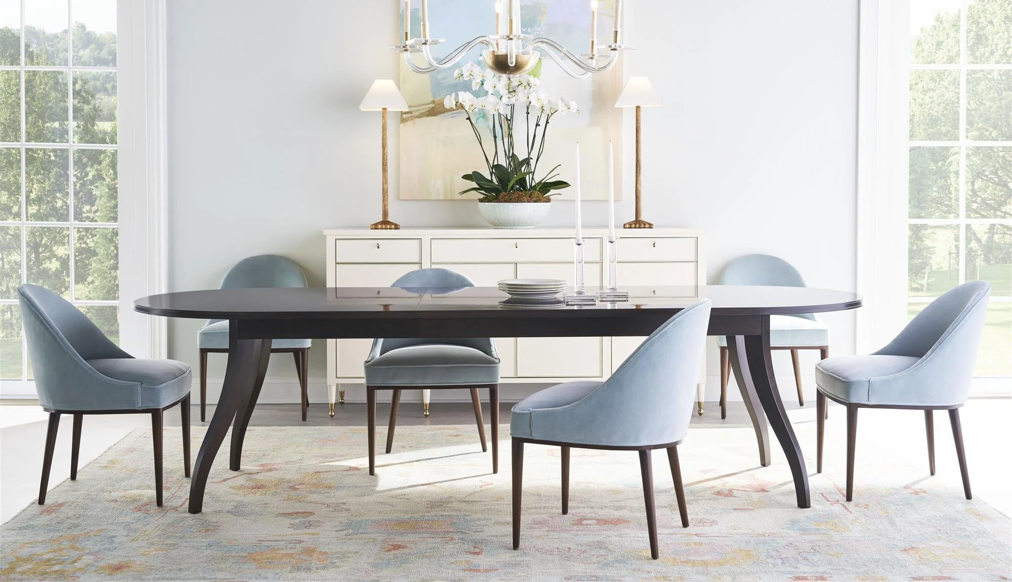 A dining room with a dark wood oval table surrounded by eight light gray upholstered chairs with dark wooden legs. The room has large windows on either side, with views of greenery outside. On the wall behind the table, there is a white sideboard wit