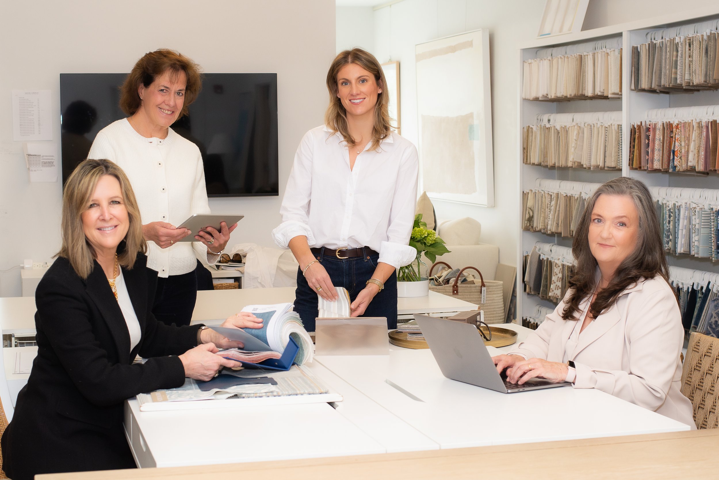 Group of women in a meeting room with fabric samples and a large shelving unit filled with fabric swatches.