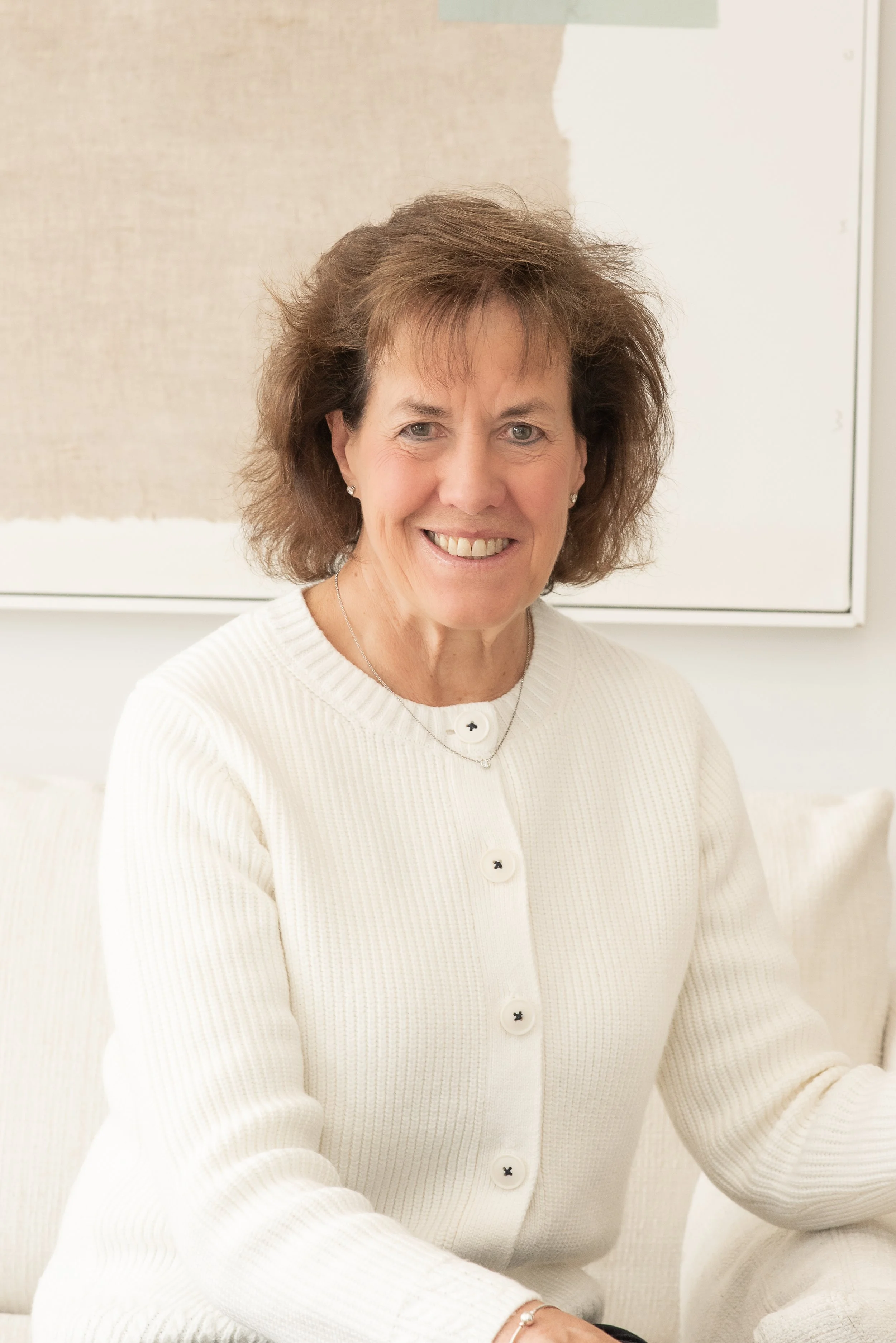 A smiling woman with curly brown hair wearing a white cardigan with black buttons, earrings, a necklace, and a ring, sitting indoors against a beige and white background.