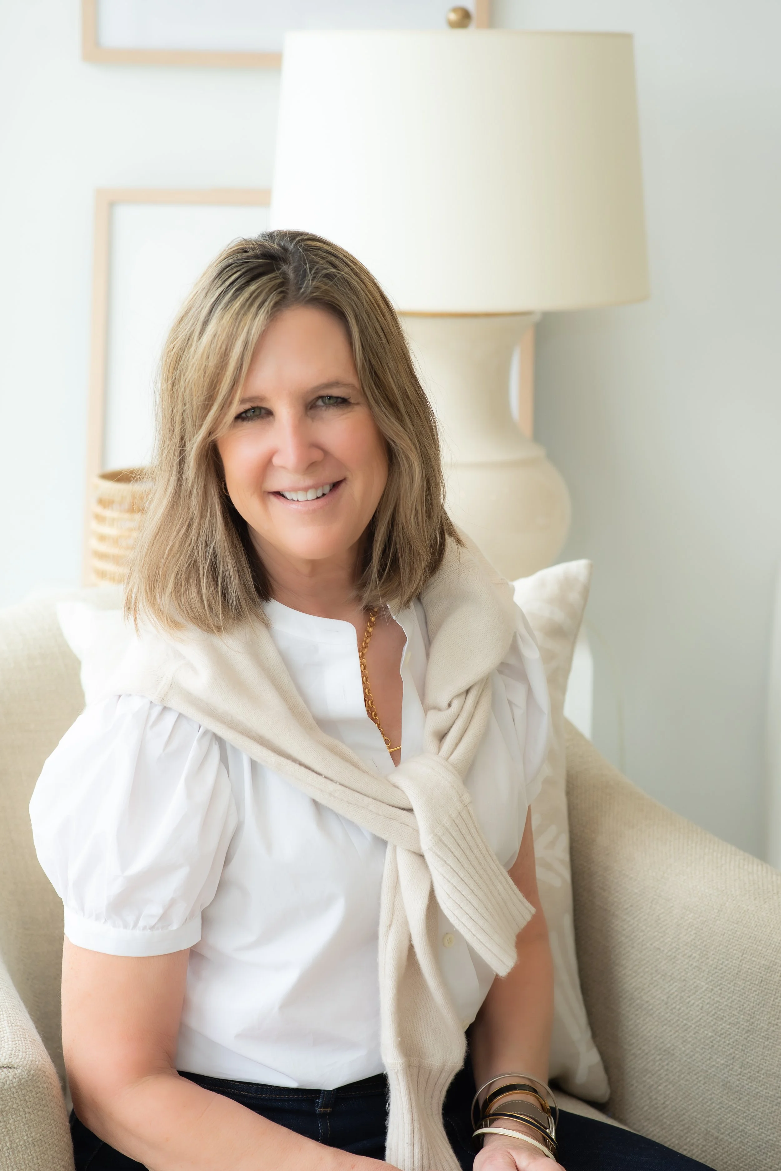 A woman with shoulder-length light brown hair smiling at the camera, sitting on a beige couch in a light-colored room with a large white lamp and picture frames on the wall behind her.