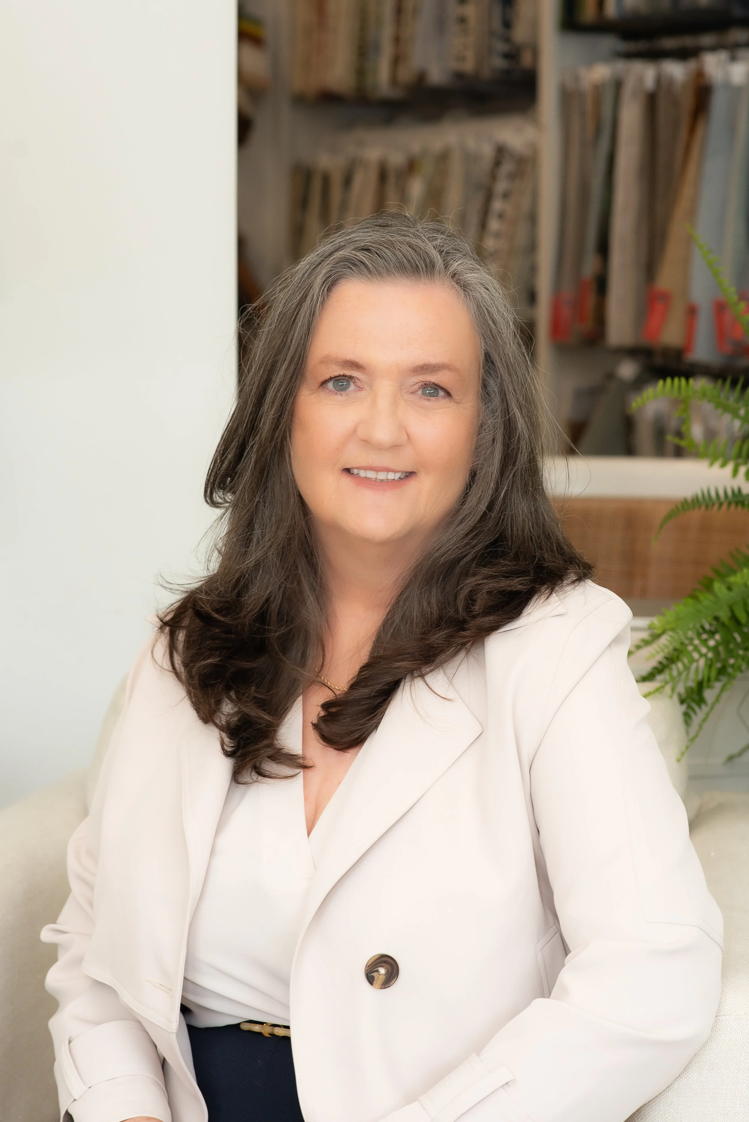 A woman with long, dark hair and blue eyes, wearing a white blazer with a button, sitting on a cream-colored chair with a bookshelf and plants in the background.