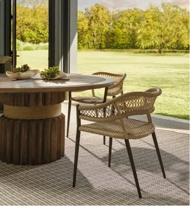 Interior view of a dining area with a large wooden table and rattan chairs, overlooking a grassy yard through large sliding glass doors.