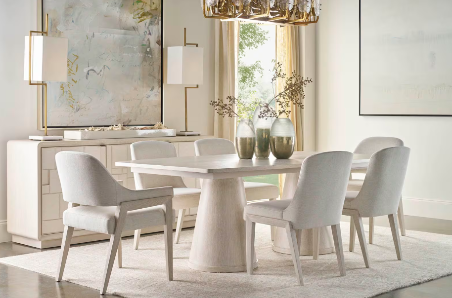 A bright, modern dining room with a white oval table surrounded by six light gray upholstered chairs. A white sideboard with decorative lamps is against the wall, and two vases with branches are on the table. Large windows with cream curtains let in 