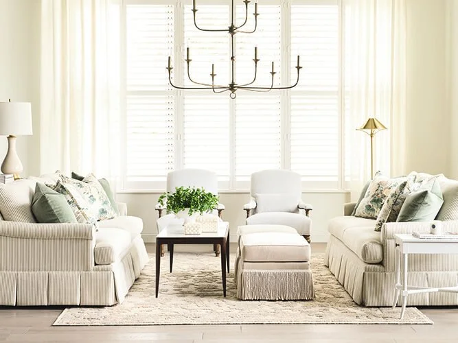 Bright living room with two white sofas, decorative pillows, a coffee table with a plant, two white chairs, a beige ottoman, a floor lamp, and a black chandelier, with large windows and white shutters.