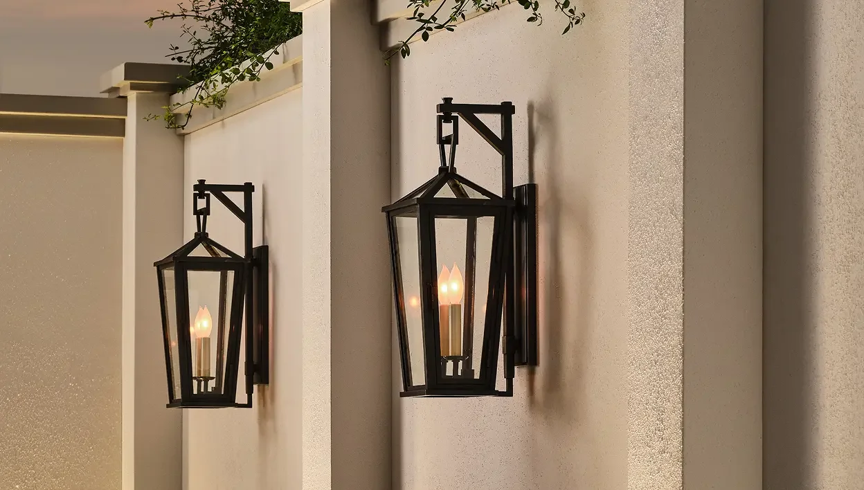 Two black wall-mounted lanterns with light bulbs against a beige wall, with greenery visible at the top.