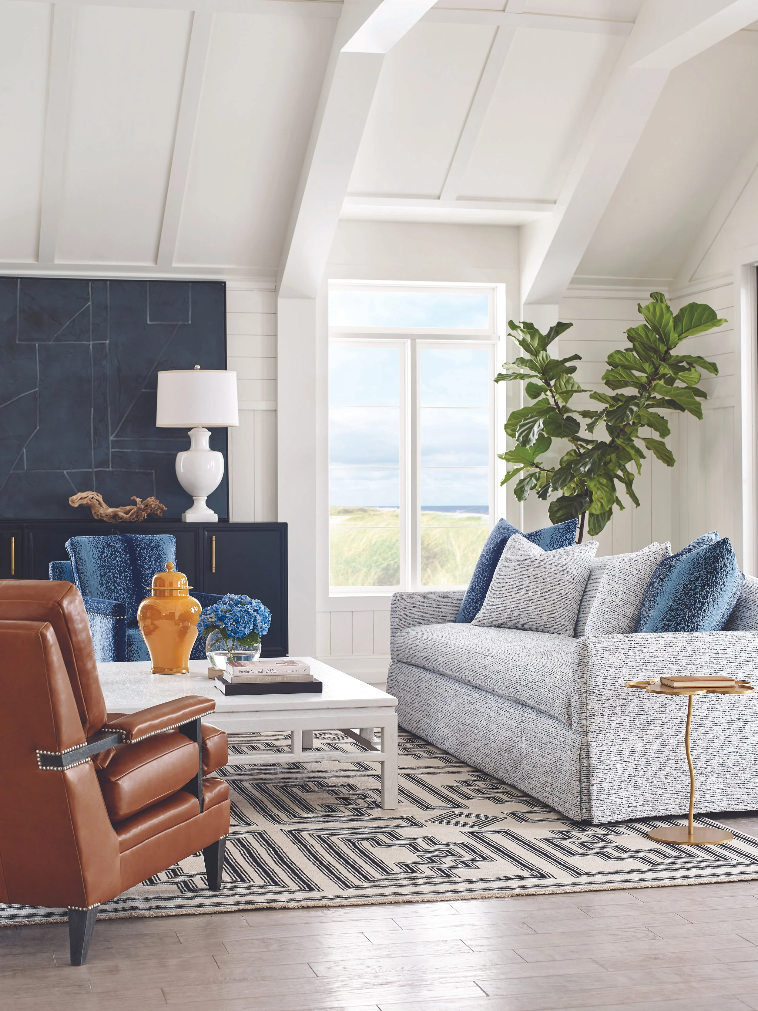 Living room with white walls, vaulted ceiling with beams, large window, a grey sofa with pillows, a large green indoor plant, a white coffee table with a blue floral arrangement, a striped rug, and a black cabinet with decor items and a white lamp.