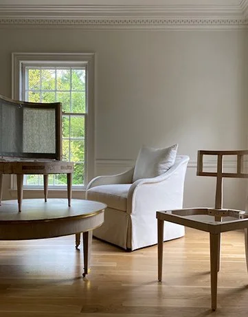 A white armchair and a wooden chair with a cardboard frame in a bright room with hardwood floors and a large window.