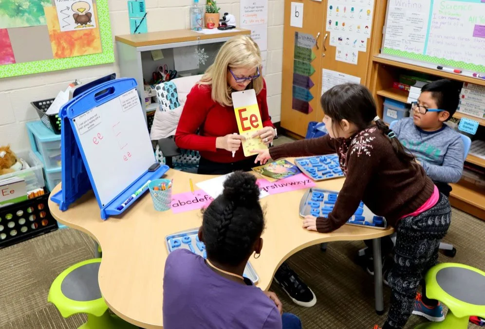A teacher sitting at a small table with four students in a classroom. The teacher is holding a large card with the letter 'E' on it, and one student is passing a book to her. On the table are phonics boards with the alphabet, markers, and a container with pencils. The classroom has educational posters and supplies on the walls and shelves.