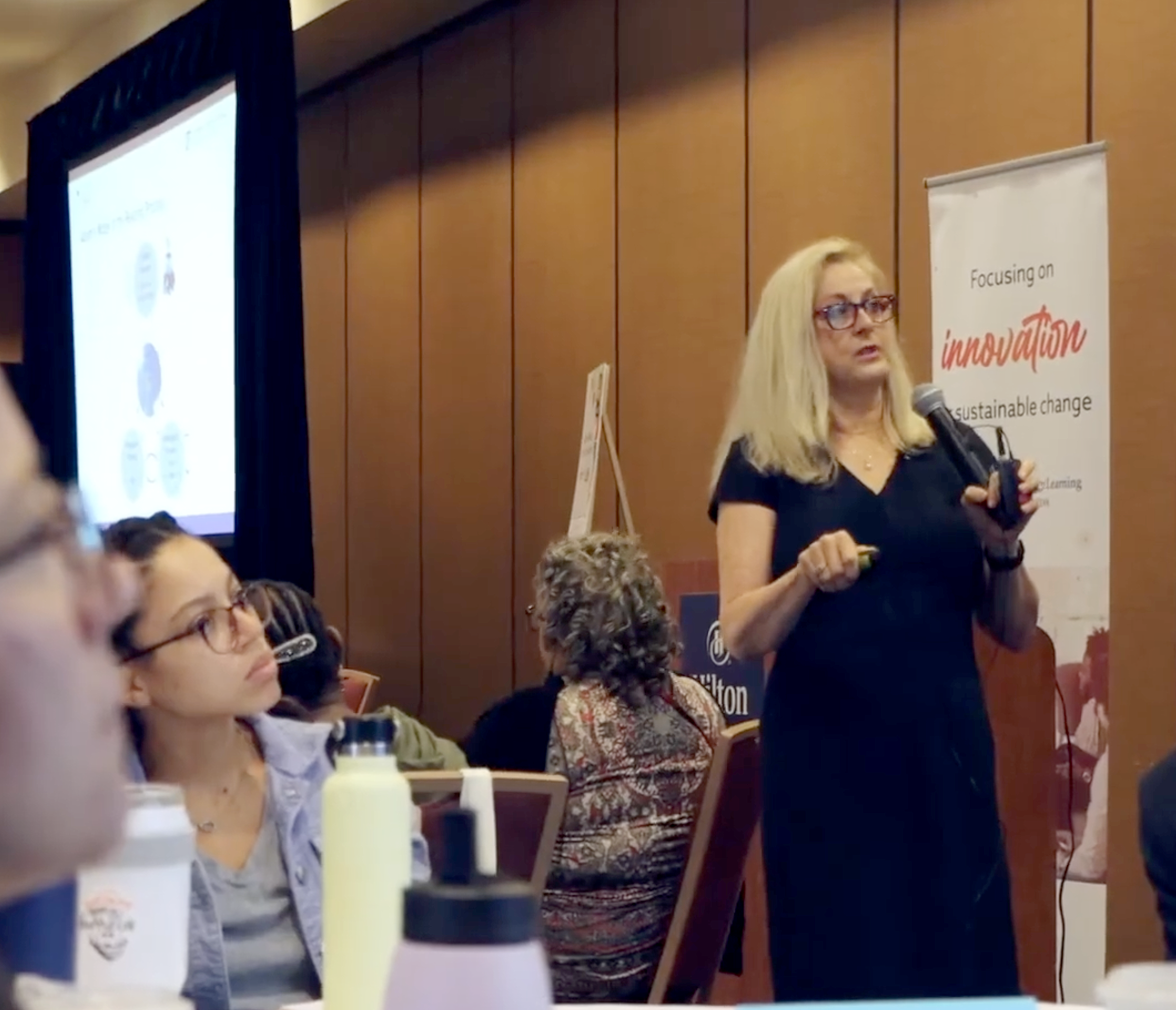 Dr. Paige is speaking into a microphone  a presentation at a conference. She is standing in front of a banner that says 'Focusing on innovation and sustainable change.' Several audience members are seated at tables, listening, with some taking notes