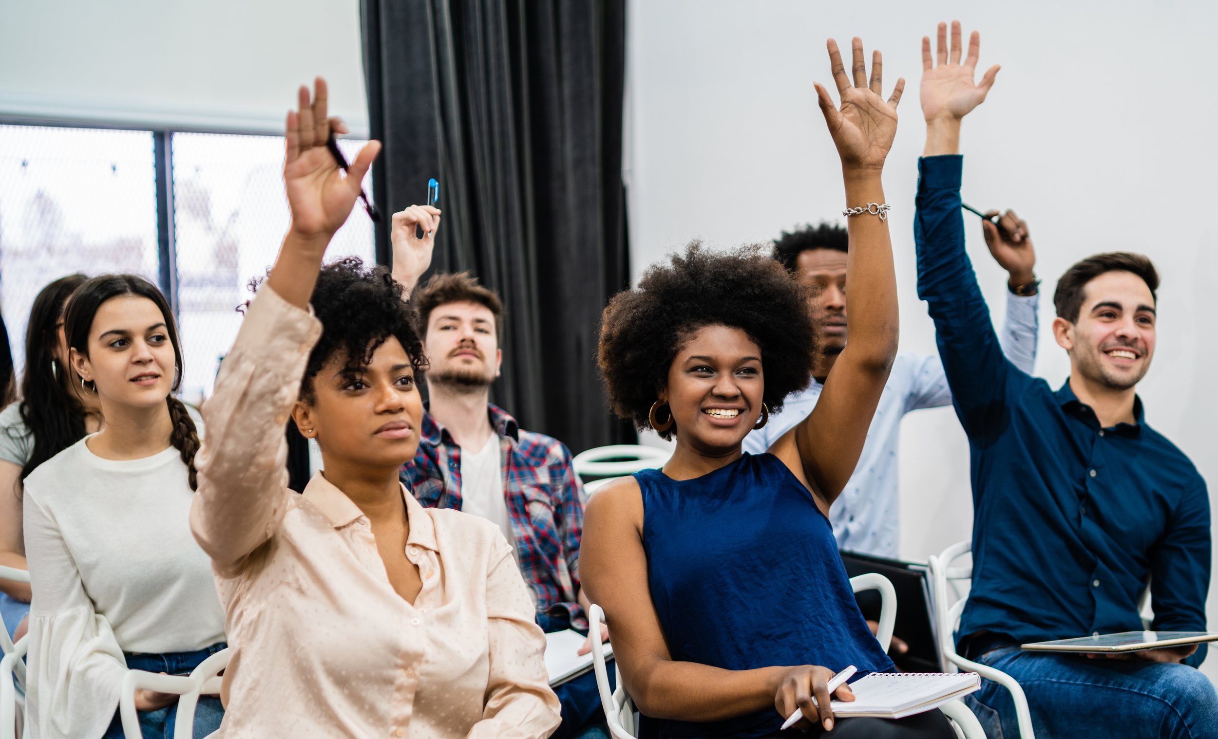 Group of diverse young teachers in a classroom raising their hands during a lecture or discussion.