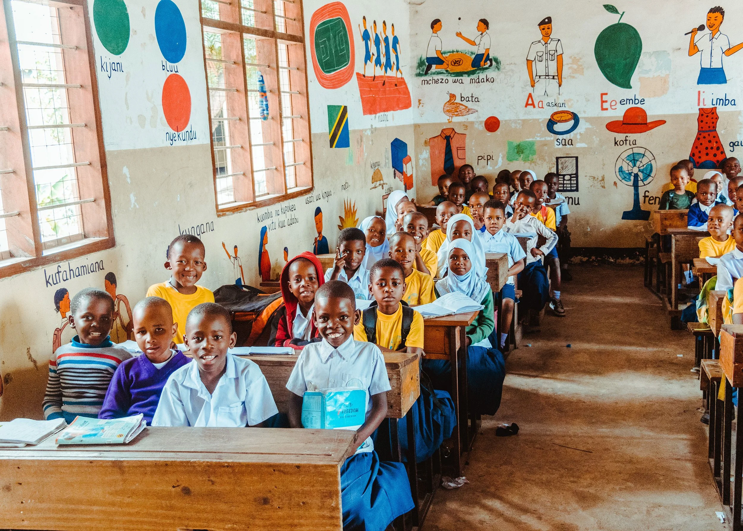 A classroom filled with young students sitting at wooden desks, smiling at the camera. The classroom walls are decorated with educational posters and colorful drawings, including animals, objects, and Swahili words. Some students are wearing white shirts and blue skirts or pants, others are in yellow shirts, with a few wearing headscarves.