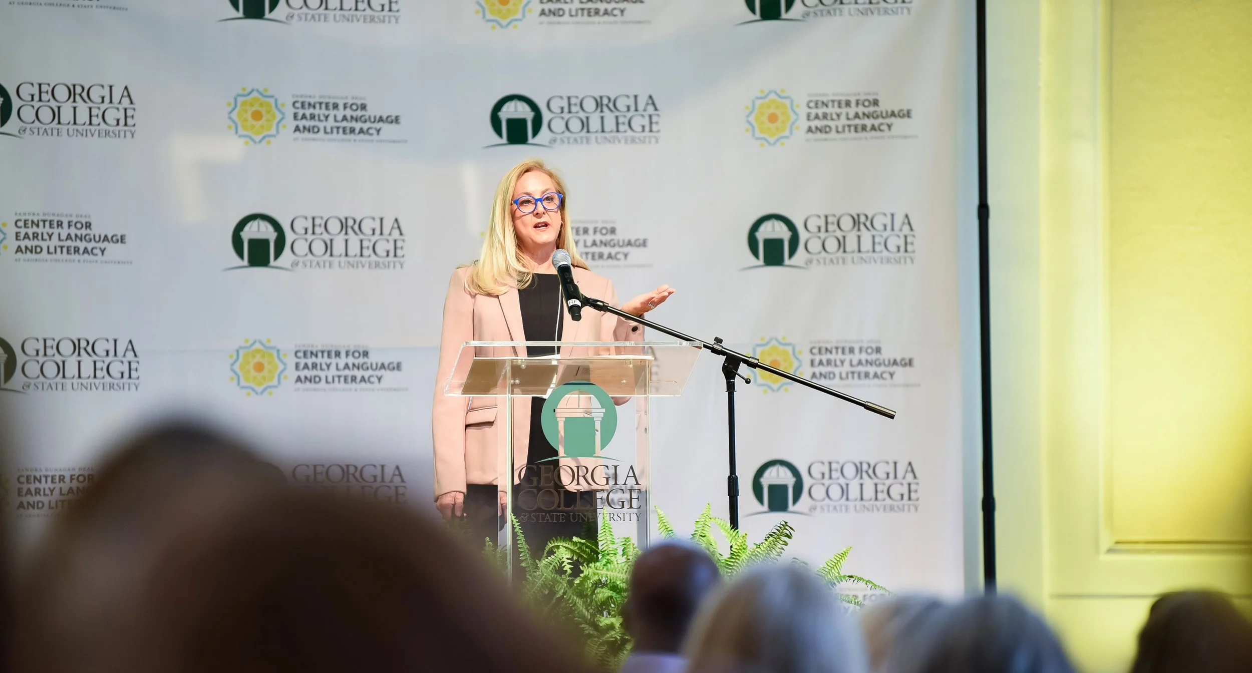A woman with blonde hair and glasses speaking at a podium during an event at Georgia College, with a backdrop displaying the college's logo and the Center for Early Language and Literacy. She is wearing a pink blazer over a black top.