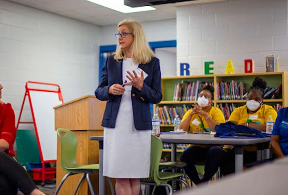 A woman with blonde hair and glasses speaking in a classroom with students sitting at tables, some wearing face masks, and bookshelves in the background.