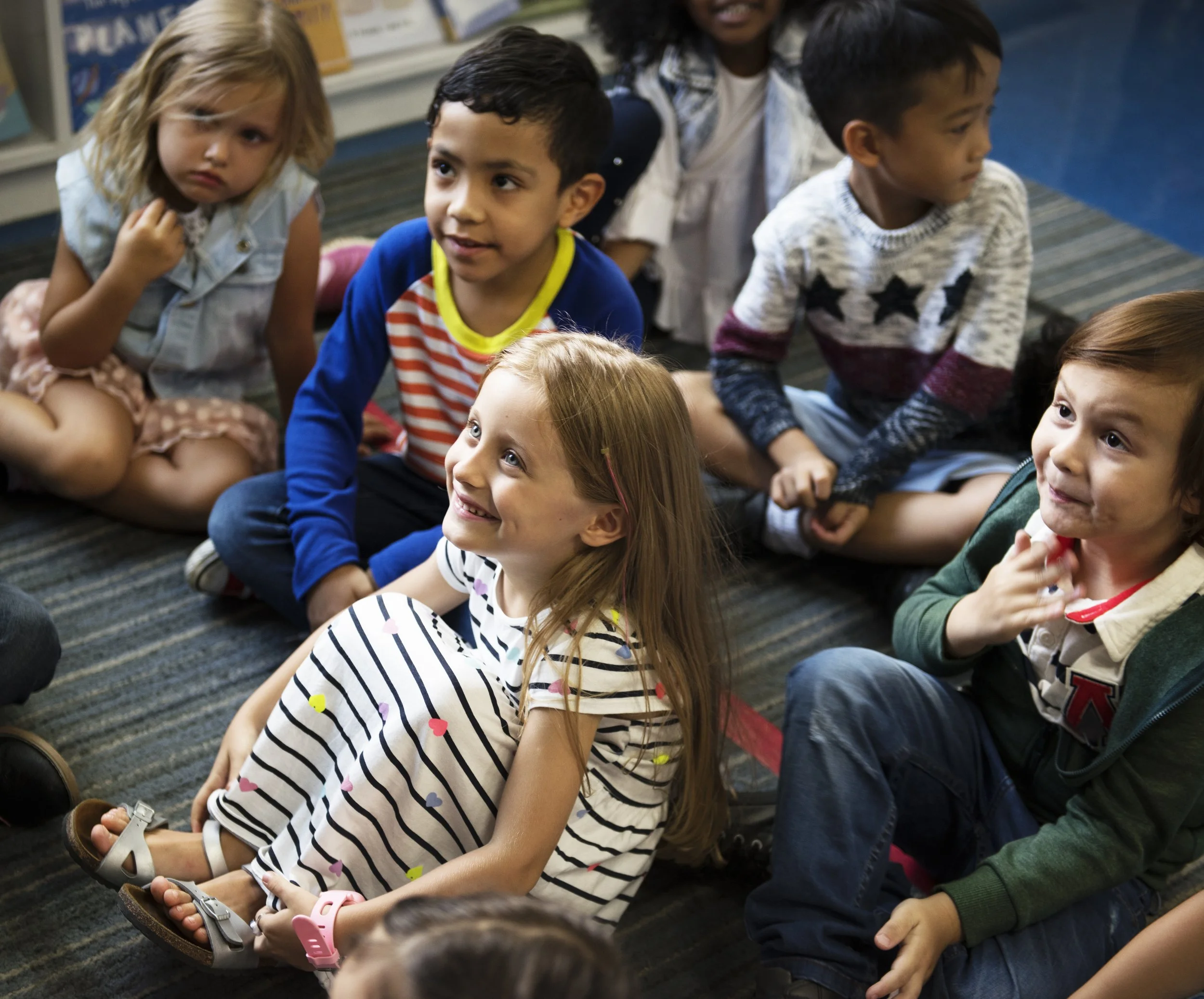 Group of young children sitting on a classroom carpet, some smiling and others looking thoughtful.