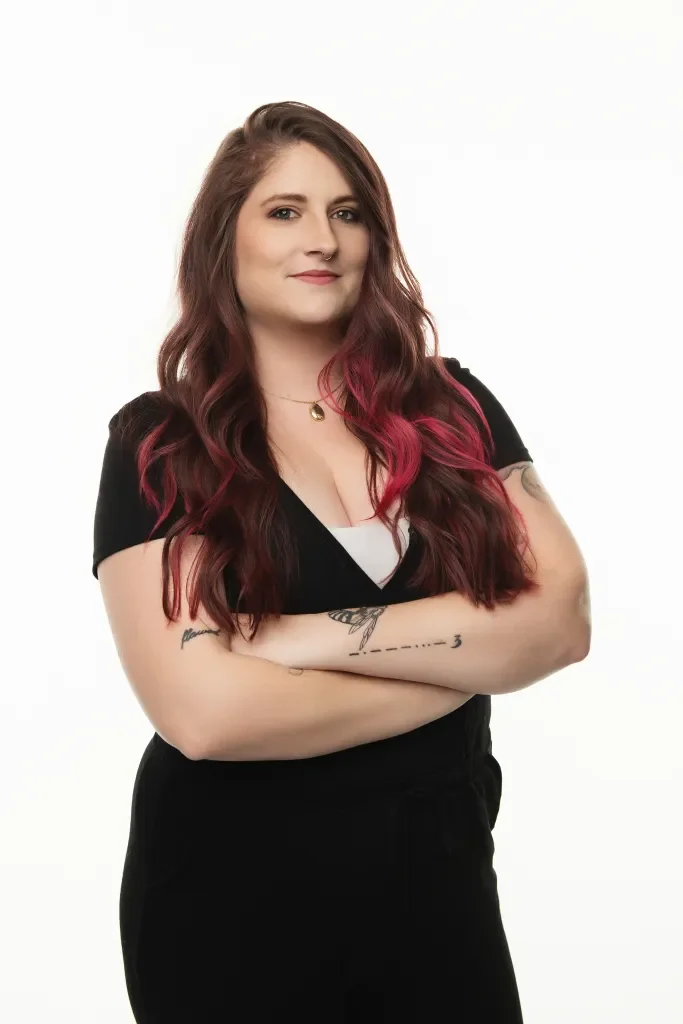 A woman with long, wavy hair dyed pink at the tips, standing against a plain white background with her arms crossed, wearing a black shirt and black pants.