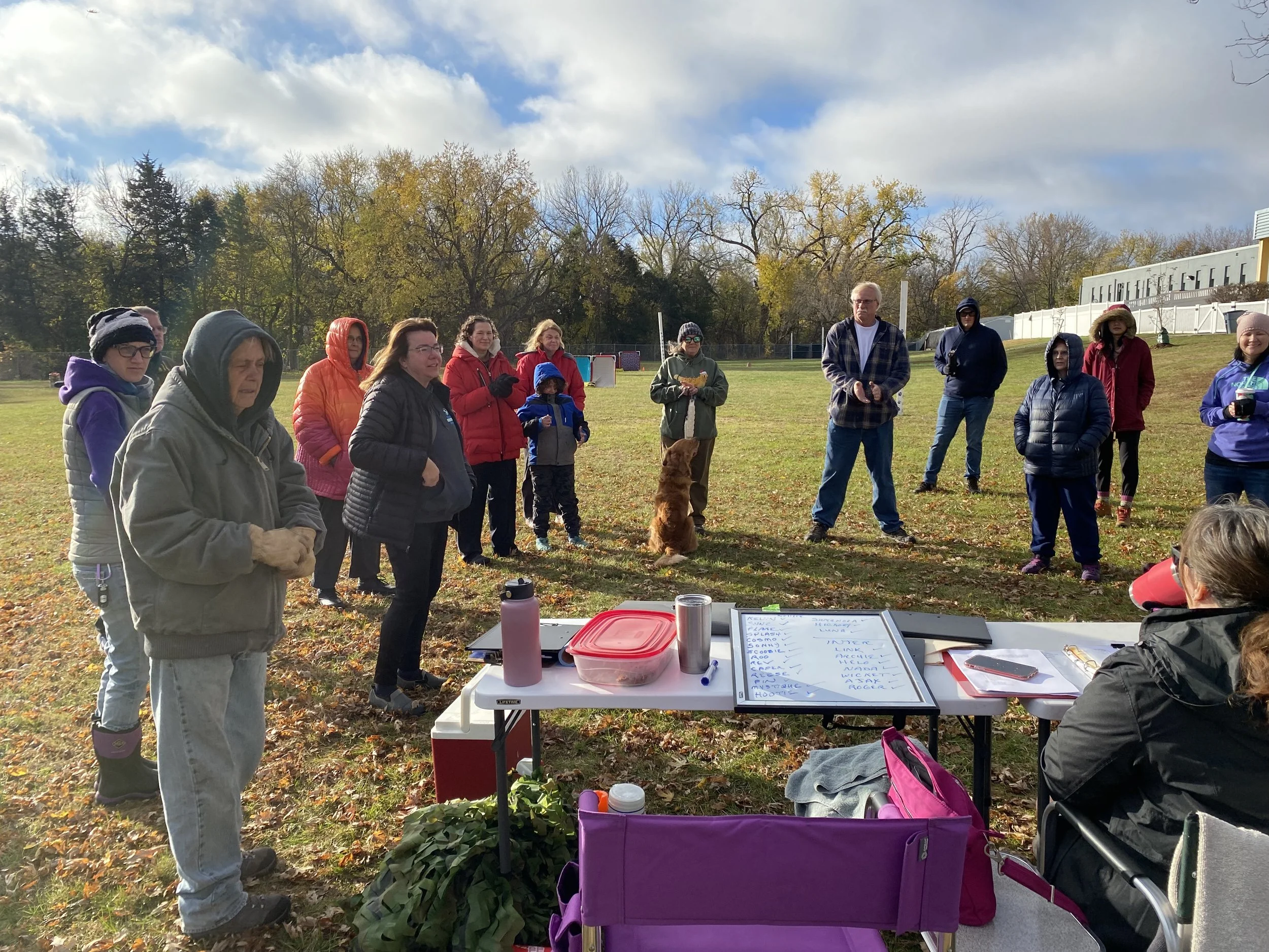 Group of people outdoors on a sunny day, standing on a grassy field, participating in an event or class, with tables and a whiteboard in the foreground.