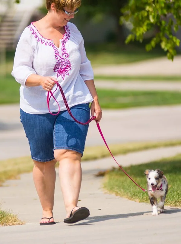 A woman walking a small dog on a pink leash on a suburban sidewalk.