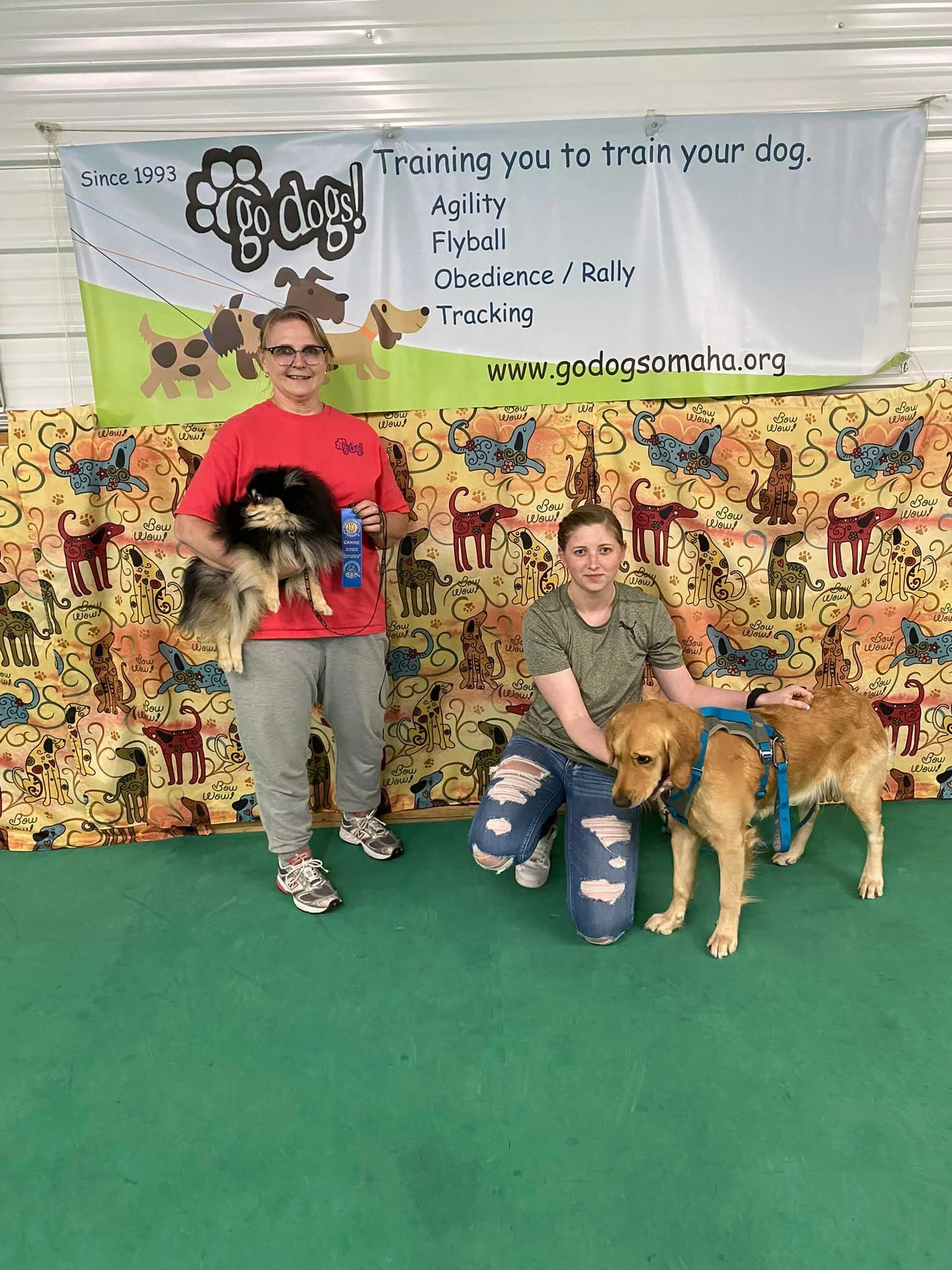 Two women with their dogs at a dog training event, standing in front of a colorful backdrop with a banner advertising dog training services. The woman on the left is holding a fluffy black and tan dog with a blue ribbon, and the woman on the right is kneeling next to a golden retriever wearing a harness.