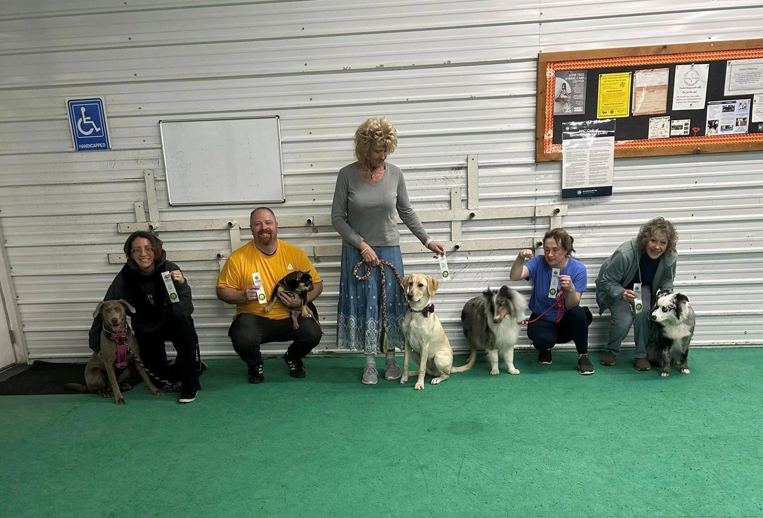Group of five people with their therapy dogs posing for a photo indoors in front of a white corrugated metal wall. The group includes three women and two men, each holding a certificate or award, with the dogs sitting or lying beside them.