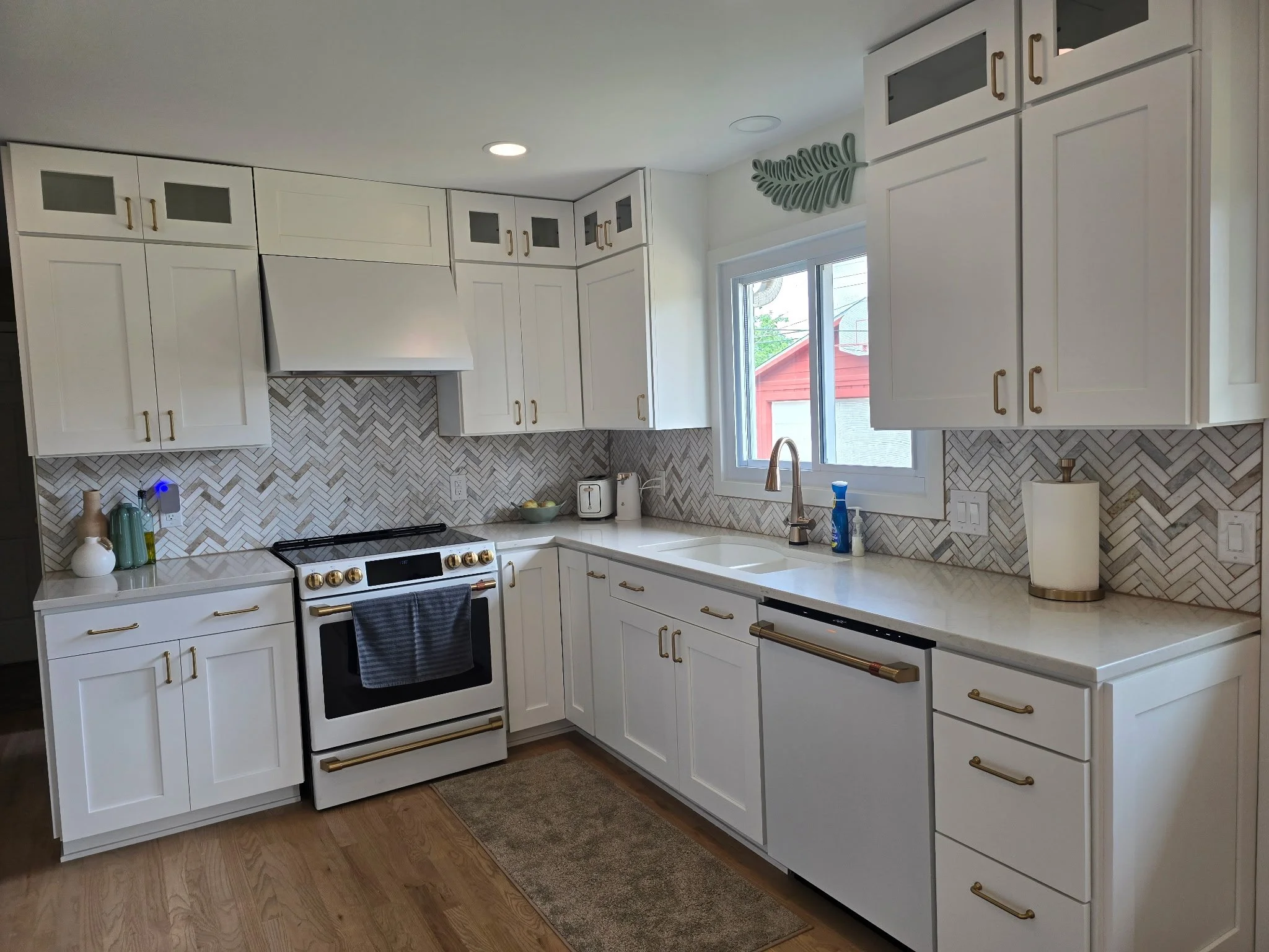 White kitchen with upper and lower cabinets, a white stove with gold handles, a window above the sink, a paper towel holder, and a patterned backsplash.