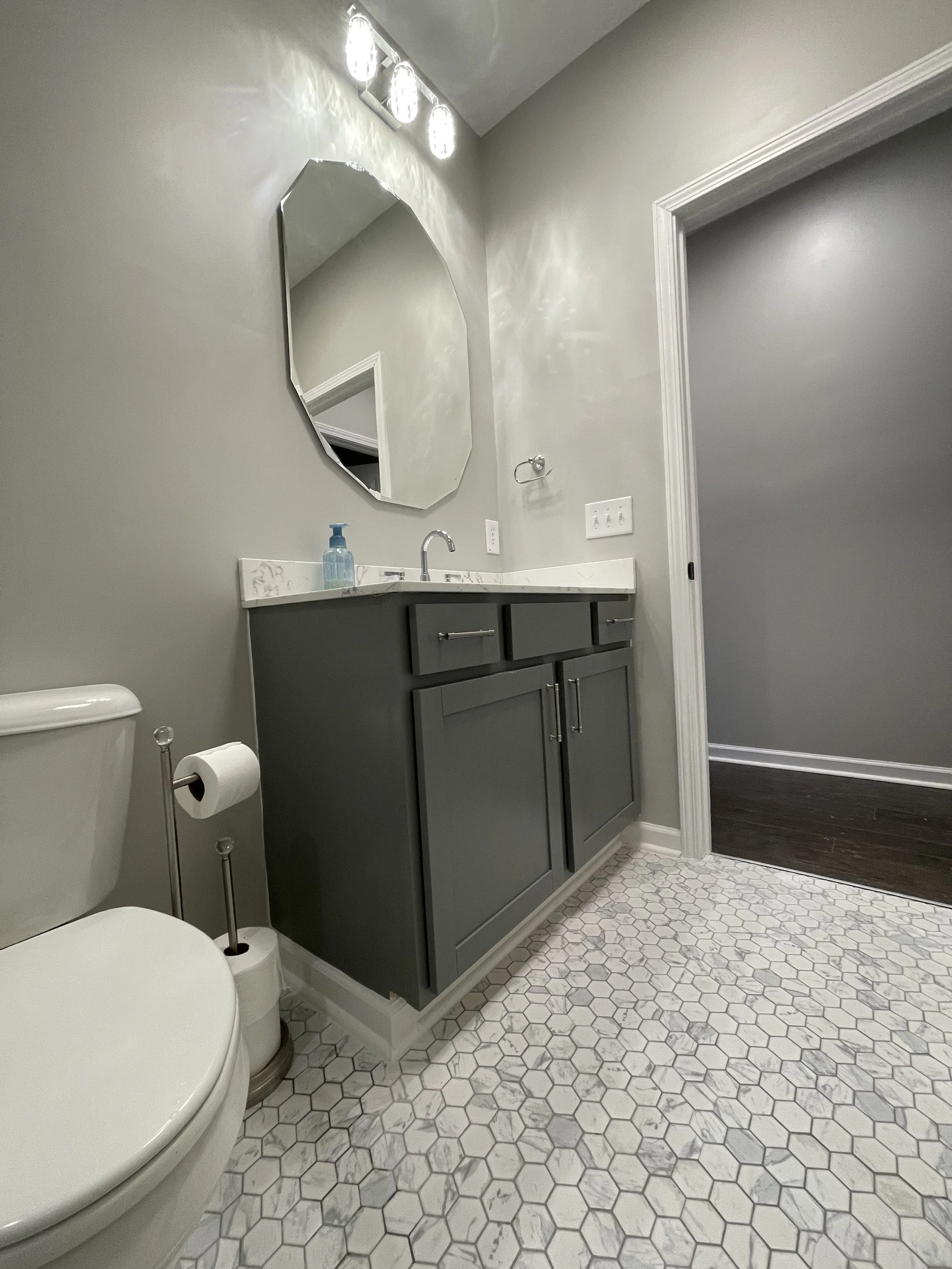 Bathroom with gray vanity, white marble countertop, oval mirror, hexagonal tile floor, toilet, blue soap dispenser, wall-mounted soap dish, and doorway leading to a room with dark hardwood floor.