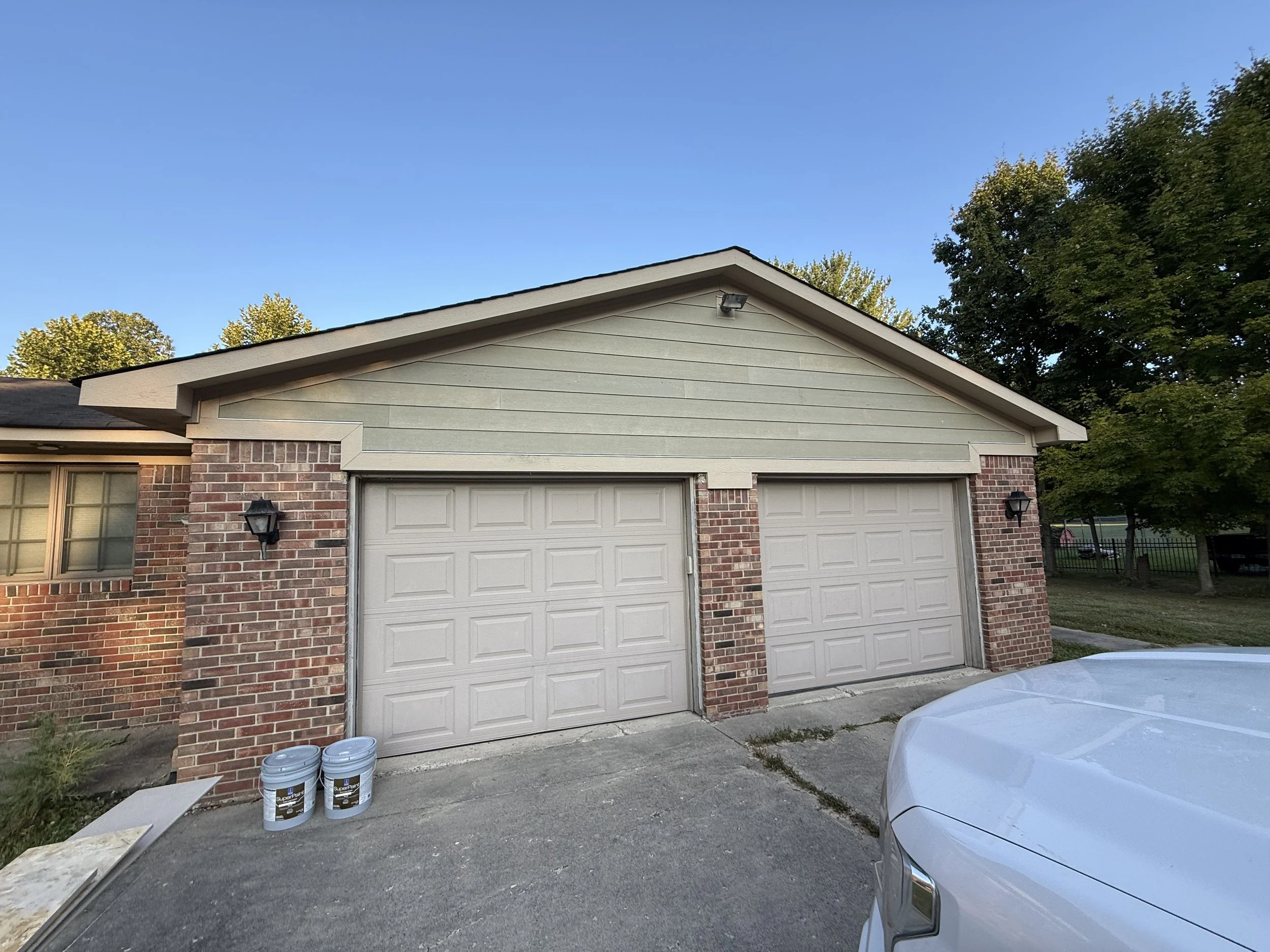 A two-car garage with brick and light green siding exterior, two garage door openers, two black wall-mounted lanterns, and a white pickup truck parked in front. Two buckets of paint are on the driveway, and trees are in the background under a clear b