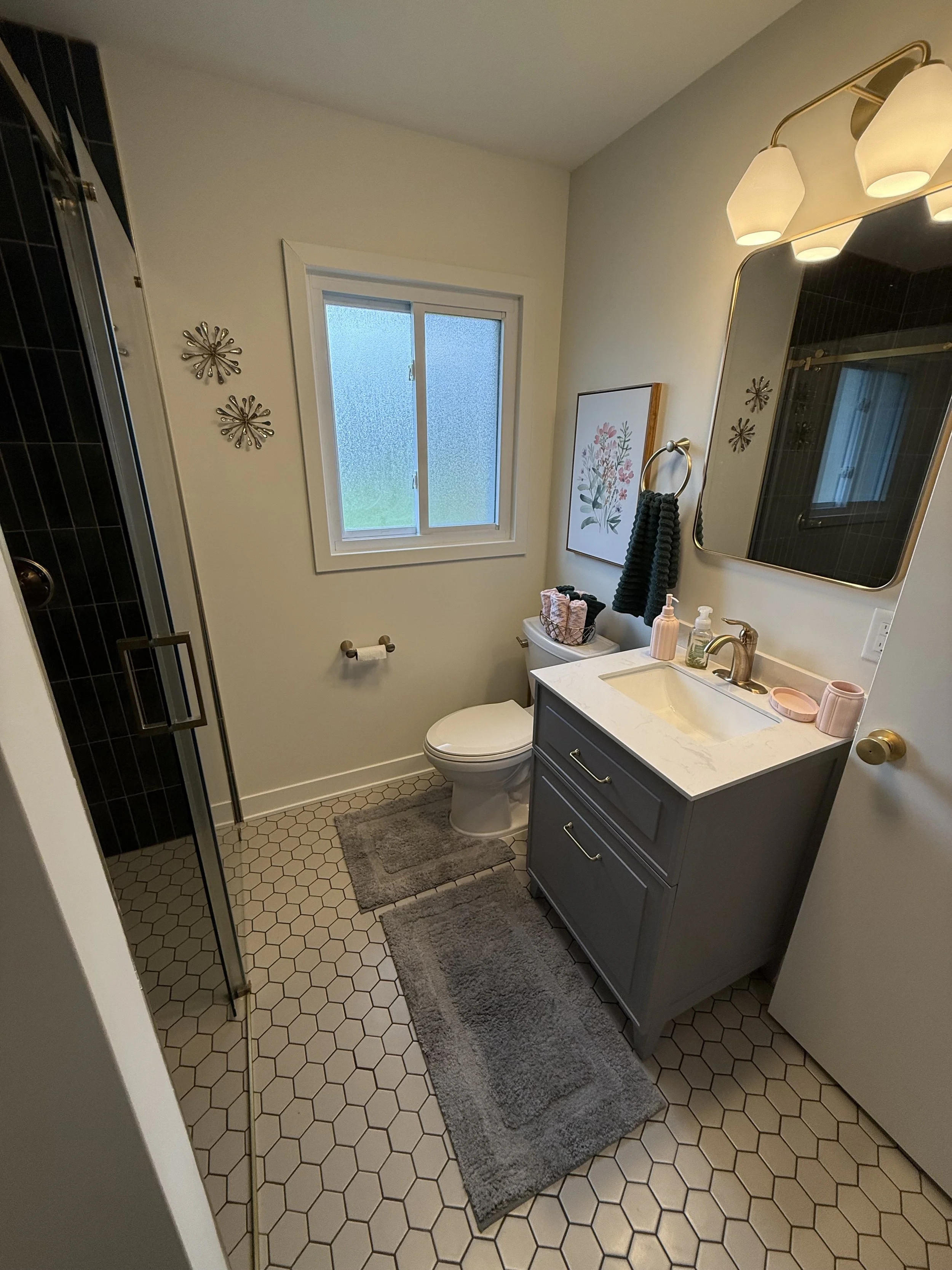 A small bathroom with hexagonal tile flooring, a gray vanity with a white countertop, a mirror with warm lighting, and a frosted window. There are two gray rugs, a toilet with a basket on top, and decorative wall art and metal wall hangings.
