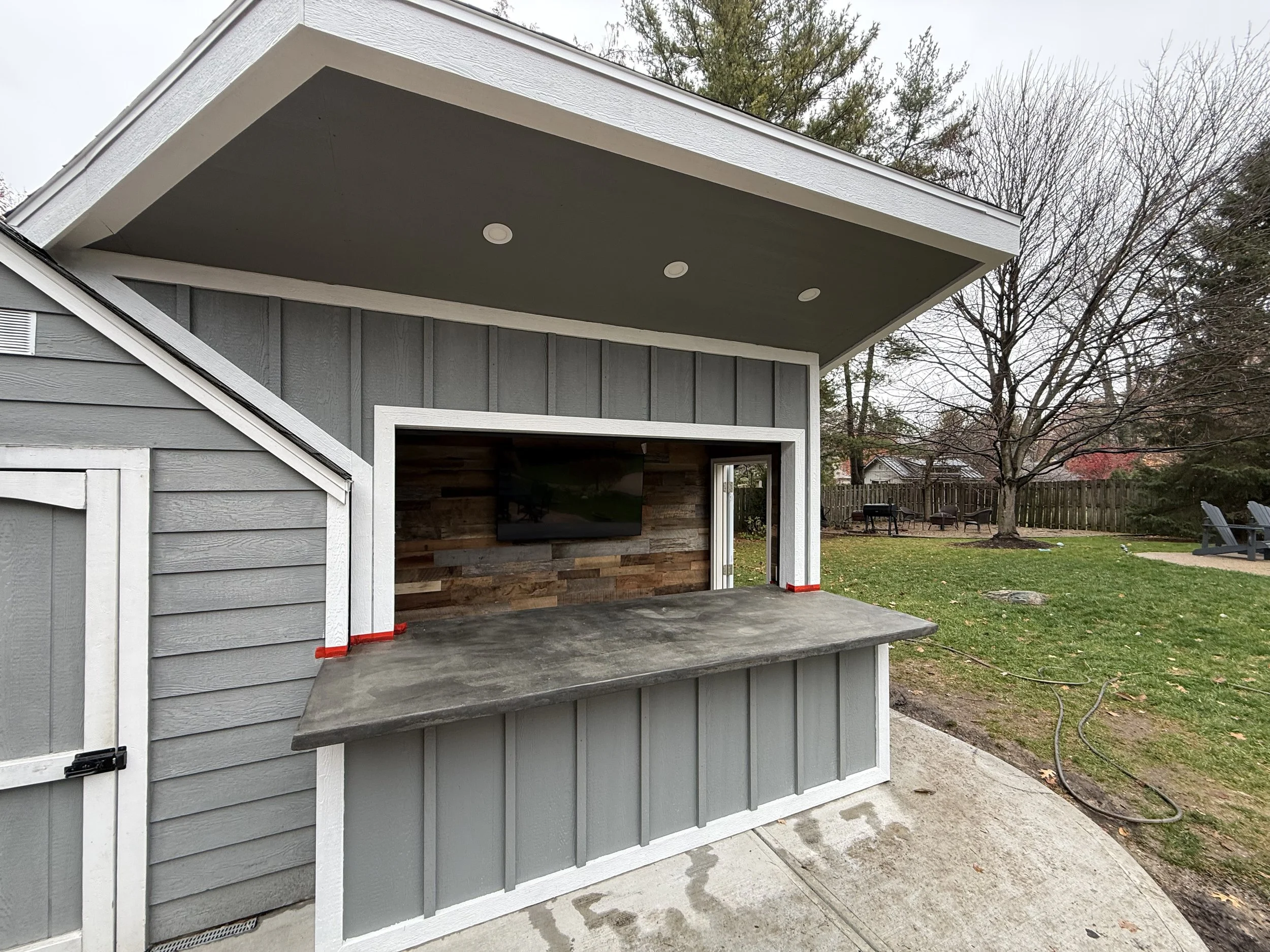 Outdoor entertainment area with a built-in TV, counter space, and a shaded roof, in a backyard with trees and lawn.