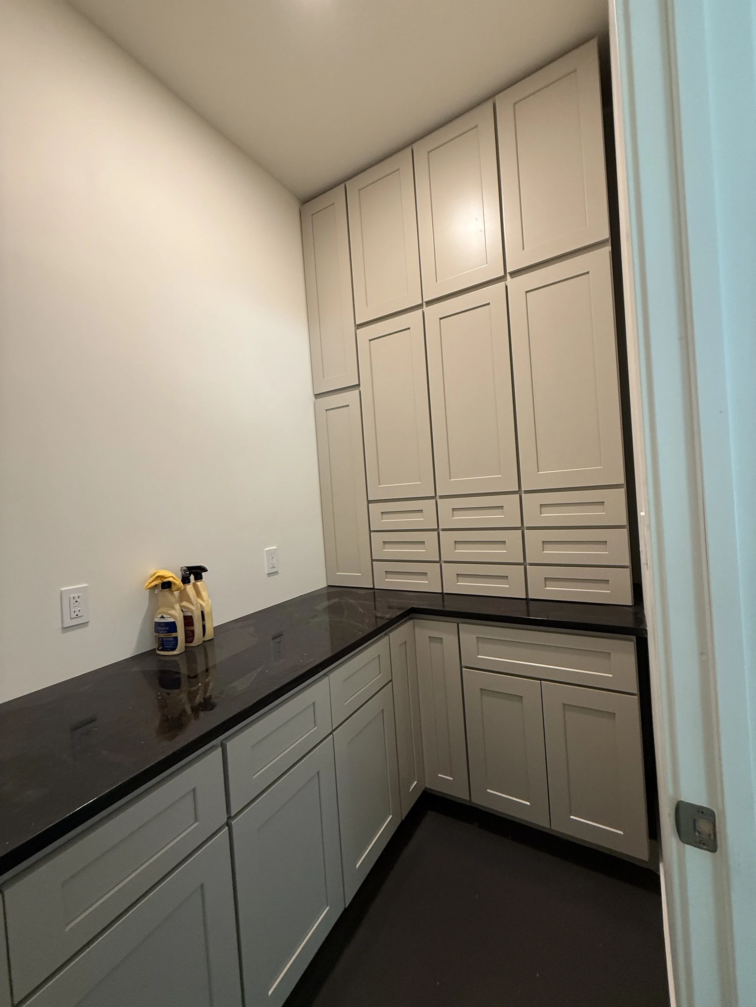 A corner of a laundry room with gray cabinets, a black countertop, cleaning spray bottles on the counter, and electrical outlets on the wall.