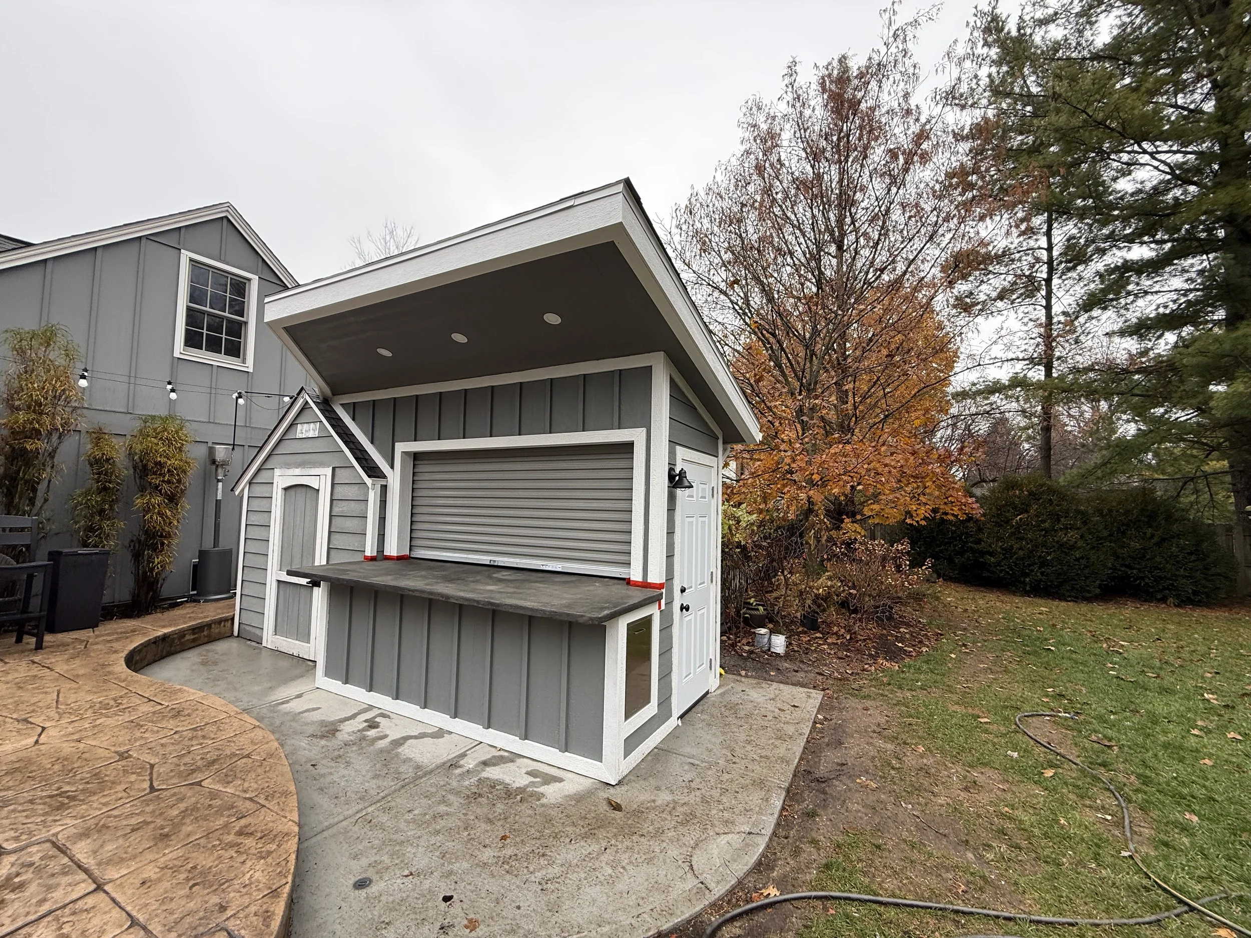 A small grey and white garden shed with a roll-up door and a mini door, situated on a concrete pad in a backyard with fallen leaves, bushes, and trees showing fall foliage.