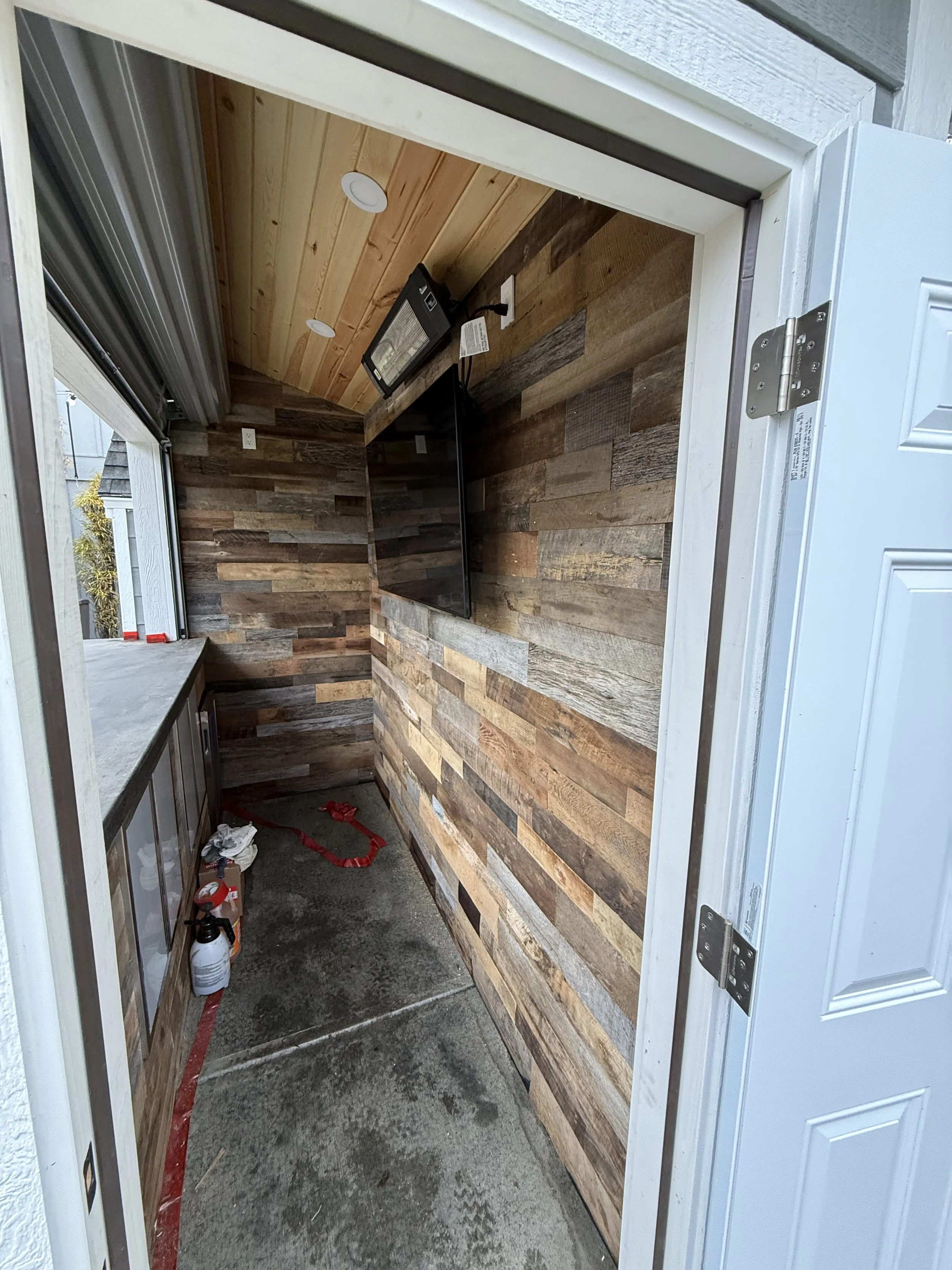 A small outdoor bar or storage area with wooden paneling on the walls and ceiling, a concrete floor, a mounted flat-screen TV, and a countertop with some tools and supplies on it.